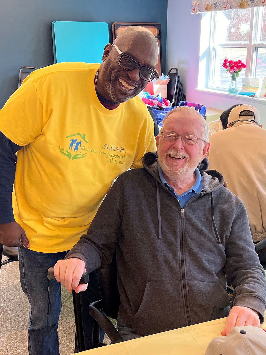 An elderly man seated with a cane smiles for a picture with a SEAH volunteer