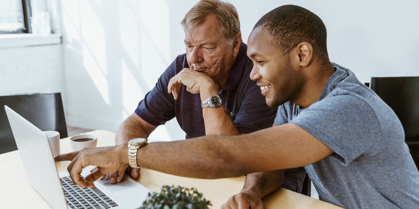 A young man points to something on the screen of his laptop as he advises an older man